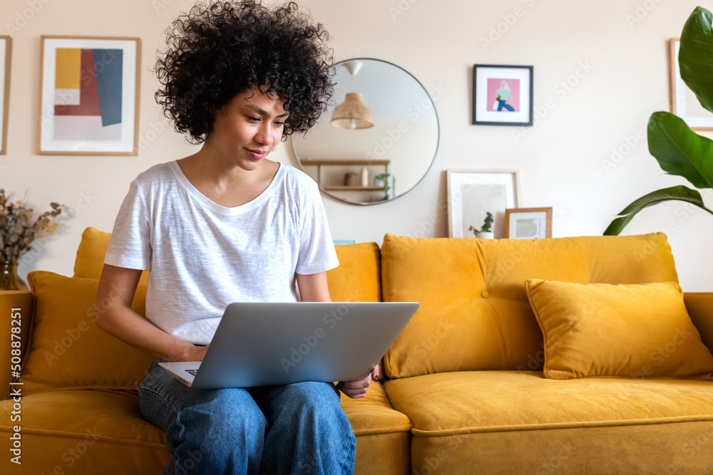 © Daniel - Young African american woman sitting on the couch using laptop working at home. Copy space. © Daniel - Young African american woman sitting on the couch using laptop working at home. Copy space.