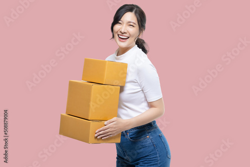 Happy Asian girl holding package parcel boxs isolated on pink screen background. Delivery courier and shipping service concept. © RedcupStudio