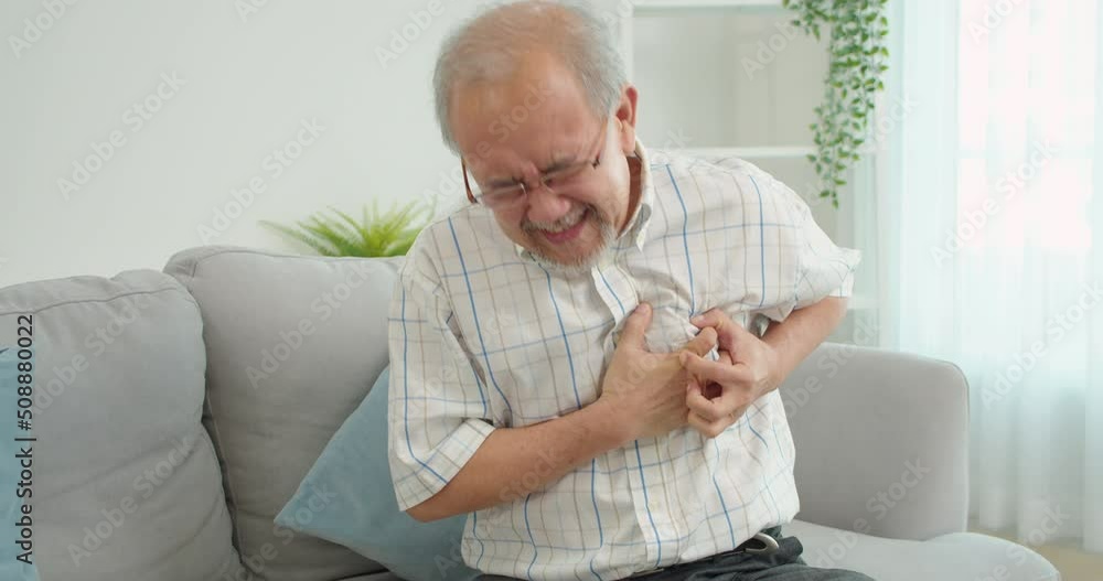 Senior male asian suffering from bad pain in his chest heart attack sitting on the sofa at home.Elderly put his hand on his left chest due to the sudden pain in his heart.Senior heart disease. Senior male asian suffering from bad pain in his chest heart attack sitting on the sofa at home.Elderly put his hand on his left chest due to the sudden pain in his heart.Senior heart disease.