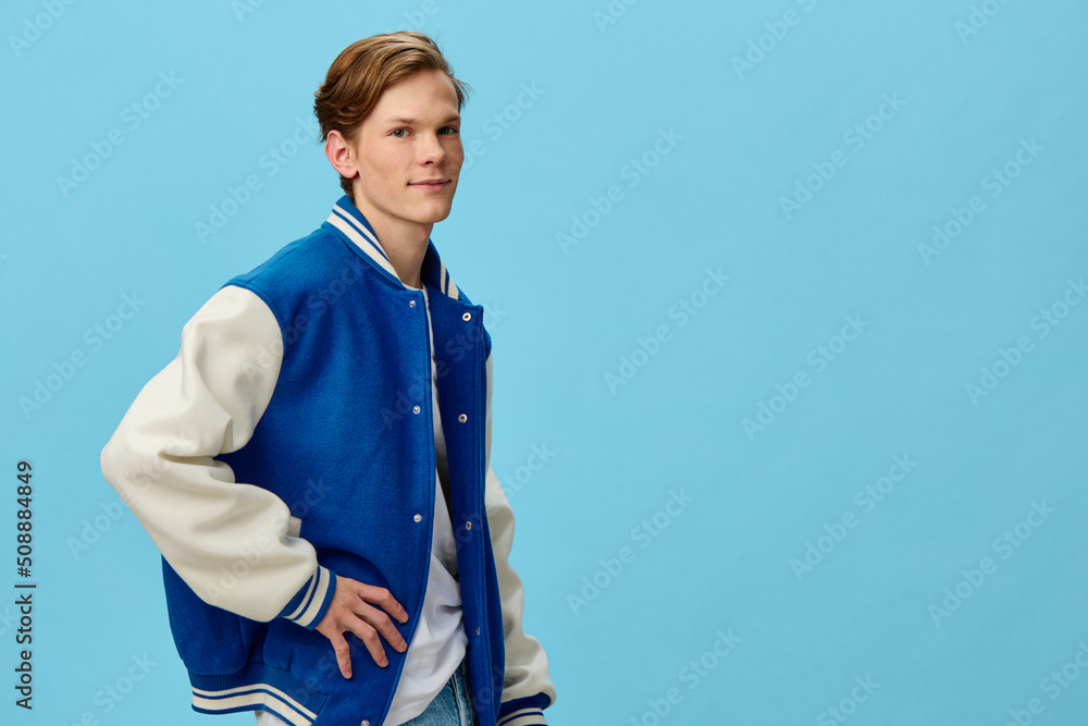 beautiful, happy an attractive young man student in a trendy bomber jacket and jeans stands with his hand on his belt on a blue background. Horizontal photo with empty space for advertising text beautiful, happy an attractive young man student in a trendy bomber jacket and jeans stands with his hand on his belt on a blue background. Horizontal photo with empty space for advertising text