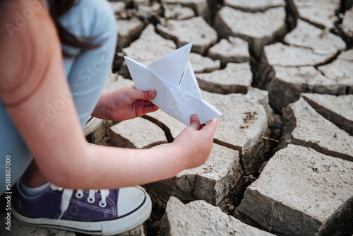 The girl lowers the paper boat onto the dry, cracked ground.