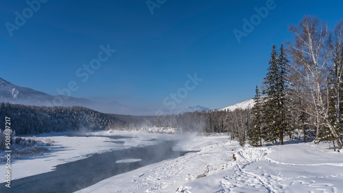 An ice-free river in a snow-covered valley. Steam rises above the water. Footprints are visible in snowdrifts, trees on the shore. Mountains against the blue sky. Altai