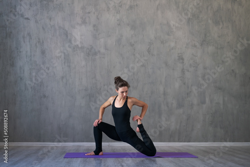 Young yogi woman practicing yoga lesson breathing meditation doing exercises and workout yoga positions, in indoors. Wellbeing, wellness concept. Girl doing stretching in sportswear against gray wall