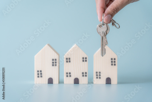 Close up of hand holding house keys selective focus at key chain small wooden house blurred at background. Blur group of wooden house at the back of keys concept of buy or rent new house.