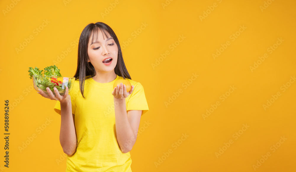 Positive cheerful slim body young woman holding glass bowl with fresh vegetables and orange in own hands while standing over isolated yellow background. Fruits and vegetables food health care concept. Positive cheerful slim body young woman holding glass bowl with fresh vegetables and orange in own hands while standing over isolated yellow background. Fruits and vegetables food health care concept.