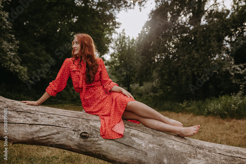 a young and beautiful woman in a red summer dress with white flecks lying on one side a fallen dry tree trunk in the middle of the forest. looking sideways