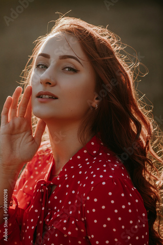 Portait of beauty young redhead woman with long curly thick hair dressed in red shirt dress with white dots posing on a camera in sunlight