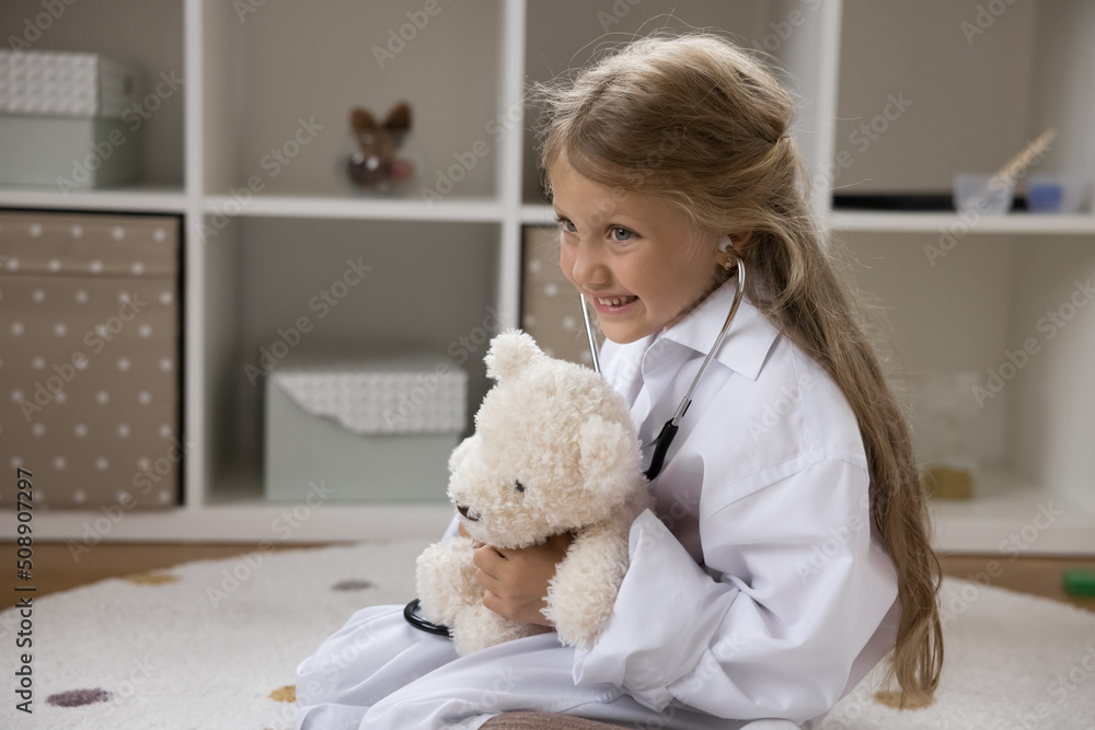 Little 6s cute girl in white coat play veterinarian use stethoscope listen fluffy toy bear heartbeat, pretend be doctor, sit on floor in nursery, make medical check up, healthcare, pediatrics concept Little 6s cute girl in white coat play veterinarian use stethoscope listen fluffy toy bear heartbeat, pretend be doctor, sit on floor in nursery, make medical check up, healthcare, pediatrics concept