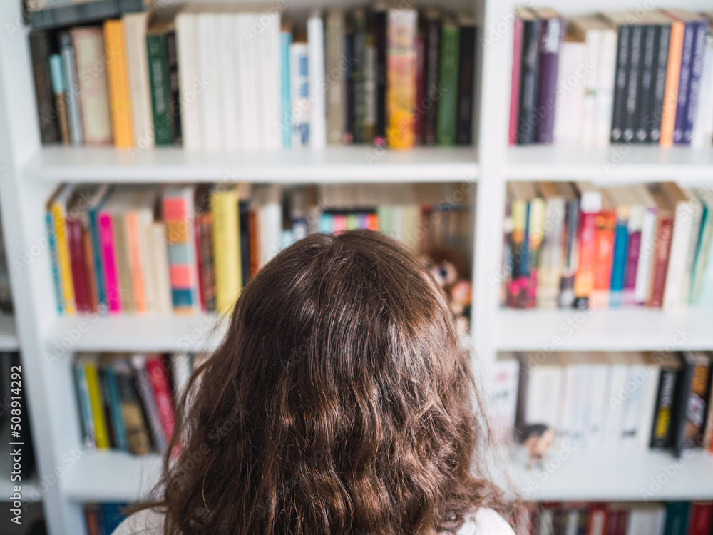 Unrecognizable woman in front of bookshelf full of books. Doubting which book to pick up in the library Unrecognizable woman in front of bookshelf full of books. Doubting which book to pick up in the library