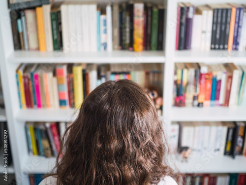 Unrecognizable woman in front of bookshelf full of books. Doubting which book to pick up in the library