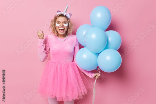 Joyful woman clenches fist celebrates special occasion feels success wears festive dress holds bunch of blue helium balloons applies beauty patches under eyes to remove wrinkles poses indoor