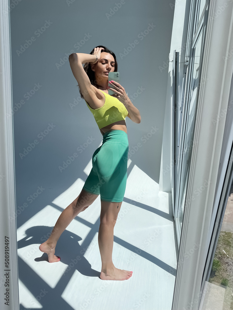 Obraz premium Fit tanned woman in sportswear, fitting blue leggings, perfect body, abs motivation, take photo selfie on phone in mirror for social media, stories, vertical.