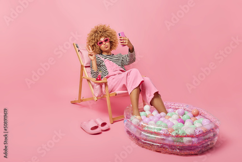 Lovely curly haired woman takes selfie makes peace gesture shares photos with friends in social networks enjoys summer time poses on deck chair keeps legs in inflated pool isolated on pink background