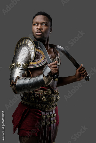 Portrait of isolated on gray gladiator of african descent dressed in light armor holding two swords.