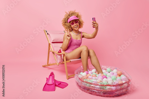 Print op canvas Indoor shot of positive woman dressed in swimwear takes selfie makes peace sign and smiles gladfully poses on deck chair isolated over pink background