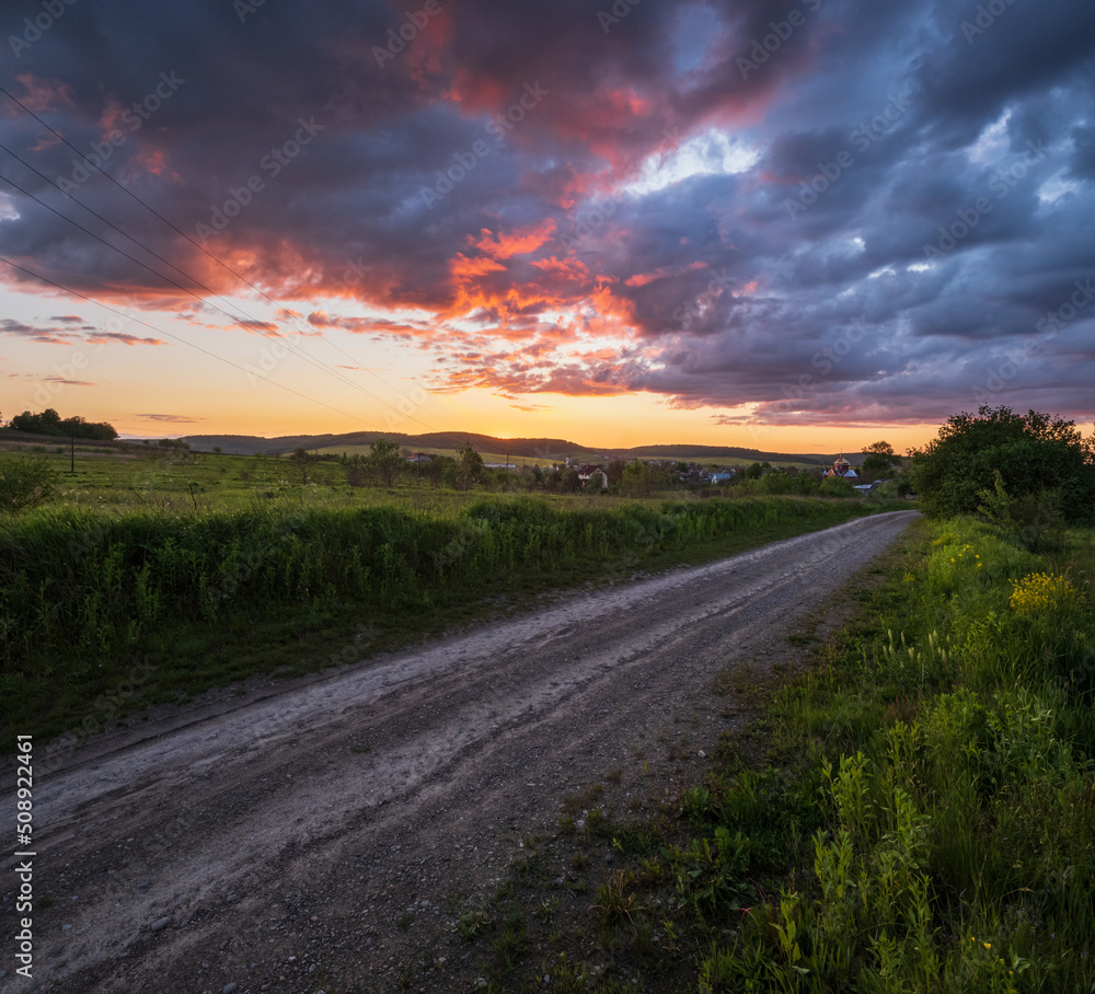 Gravel countryside road through spring meadow, cloudy evening sunset sky, rural hills, village and fields in far.