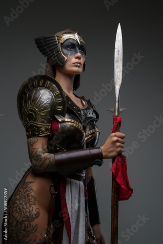 Shot of antique female warrior dressed in steel armor holding spear against grey background.