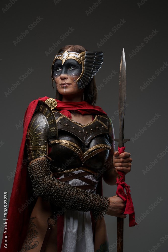 Obraz premium Studio shot of ancient woman soldier dressed in steel armor with cape holding spear.