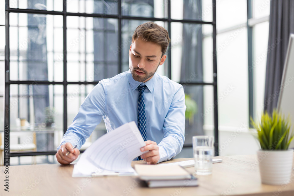 Young man is working with papers while sitting in the office. Successful entrepreneur is studying documents with attentive and concentrated look. Young man is working with papers while sitting in the office. Successful entrepreneur is studying documents with attentive and concentrated look.