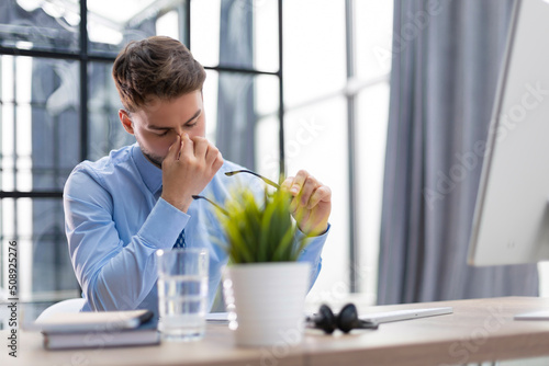 Handsome young business man with closed eyes touching face with hands while sitting on working place in office.