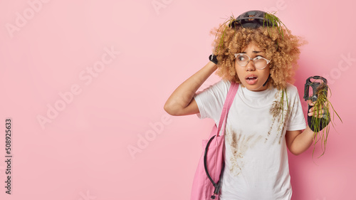 Embarrassed curly haired woman prepares for bike marathon has broken bicycle tries to solve problem carries fabric bag lock and pedal looks amazed away isolated over pink background copy space