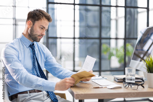 Handsome entrepreneur reading a letter from envelope in a desktop at office.