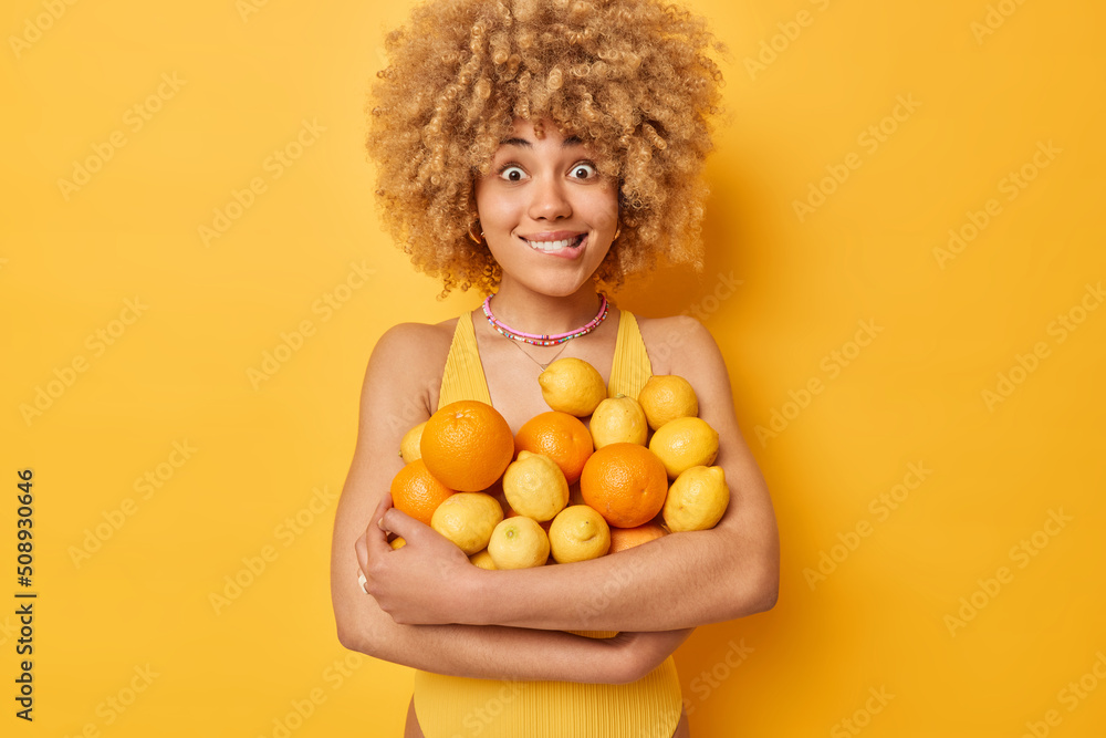 Surprised cheerful woman bites lips carries big heap of fresh oranges and lemons eats fruits full of vitamins dressed in casual t shirt isolated over vivid yellow background. People summer time - obrazy, fototapety, plakaty 