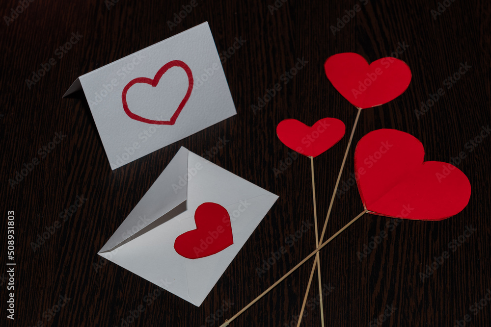 Red hearts made of cardboard, on straw stands. Red hearts and a white valentine card, with a white envelope, on a dark substrate with a woody texture.