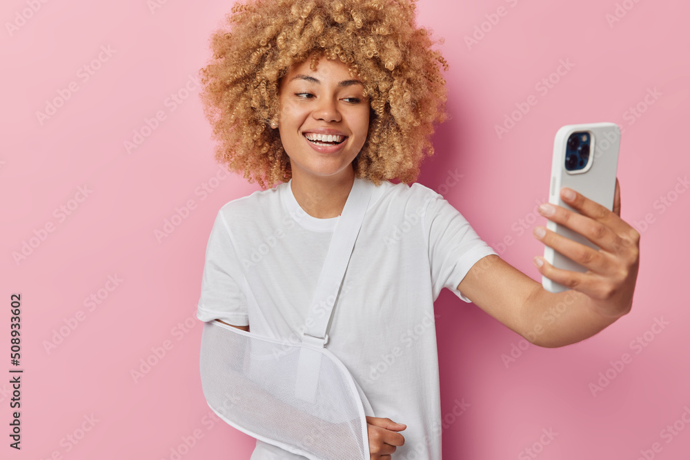 Positive woman with fair curly hair takes selfie has injured or broken arm wears splint being on her way home after visit traumatologist dressed in casual white t shirt isolated over pink background Positive woman with fair curly hair takes selfie has injured or broken arm wears splint being on her way home after visit traumatologist dressed in casual white t shirt isolated over pink background