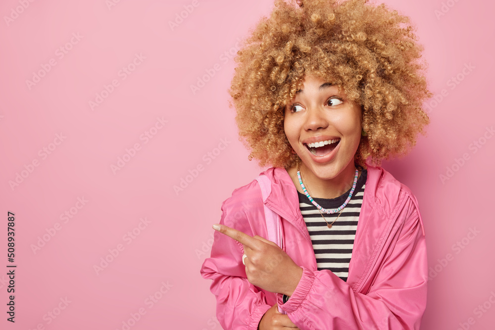 Cheerful impressed young woman with curly hair points away on blank space demonstrates awesome advertisement wears striped jumper and jacket isolated over pink background. Wow just look there