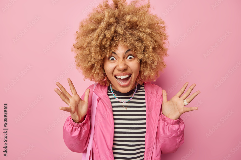 Emotional crazy curly haired woman exclaims outrages gestures angrily keeps palms raised wears casual striped jumper and jacket isolated over pink background. Human emotions and reactions concept