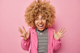 Emotional crazy curly haired woman exclaims outrages gestures angrily keeps palms raised wears casual striped jumper and jacket isolated over pink background. Human emotions and reactions concept