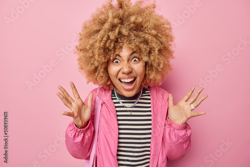 Emotional crazy curly haired woman exclaims outrages gestures angrily keeps palms raised wears casual striped jumper and jacket isolated over pink background. Human emotions and reactions concept