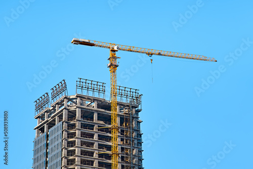 Top of the cement and glass skyscraper under construction with a yellow tower crane along it against a clear blue sky in sunny weather