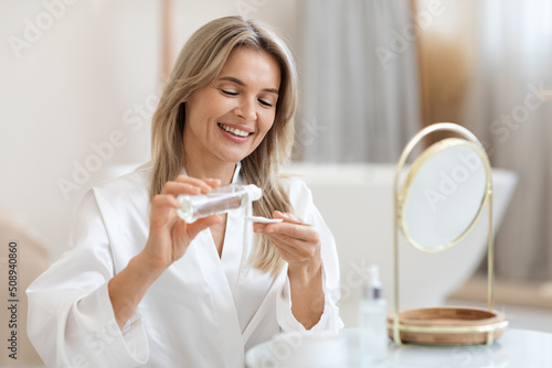 Pretty blonde woman using face tonner, looking at mirror photo