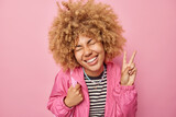 Happy carefree woman with curly hair shows peace v sign laughs gladfully keeps eyes closed dressed in jacket expresses positive emotions isolated over pink background. Body language concept.