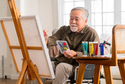 old senior asian male using art therapy while his legs is injured at home,asian elder man spend recovering of his leg is getting better with painting canvas art in living room at home