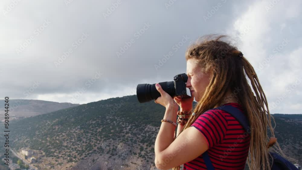 Attractive girl with dreadlocks takes pictures of nature. Profile side view. Young woman professional photographer shoot photos of landscape with mountains Attractive girl with dreadlocks takes pictures of nature. Profile side view. Young woman professional photographer shoot photos of landscape with mountains