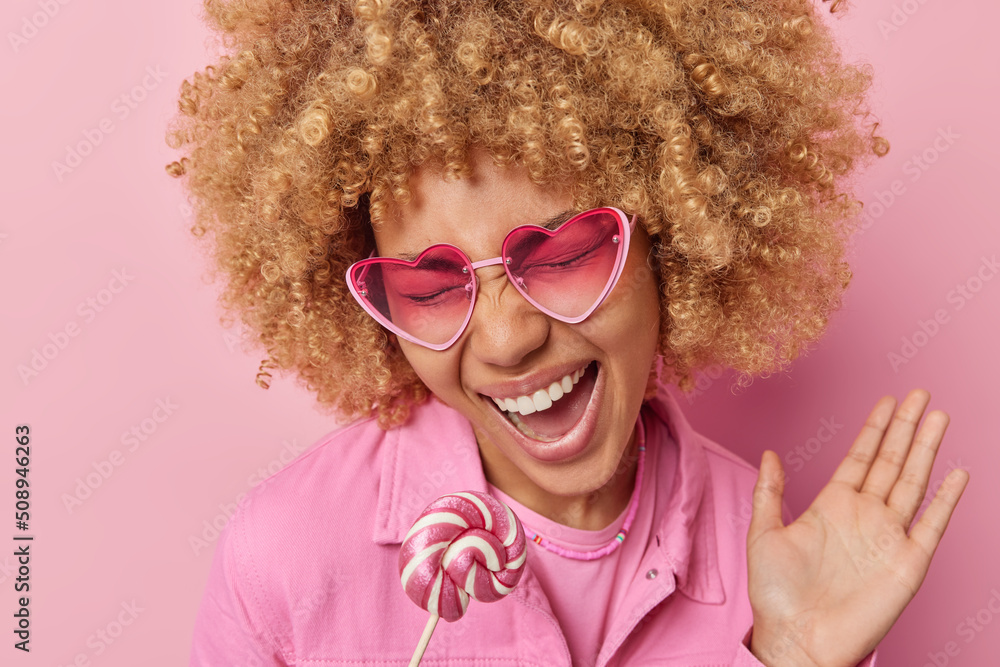 Headshot of optimistic carefree woman with curly hair exclaims loudly keeps palm raised holds lollipop near mouth has sweet tooth wears trendy sunglasses and jacket isolated over pink background. Headshot of optimistic carefree woman with curly hair exclaims loudly keeps palm raised holds lollipop near mouth has sweet tooth wears trendy sunglasses and jacket isolated over pink background.