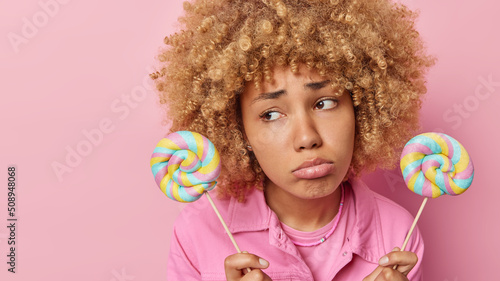 Unhappy woman with curly hair holds two colorful lollipops has sad expression keeps to diet purses lips wears fashionable jacket isolated over pink background empty space for your promotion.
