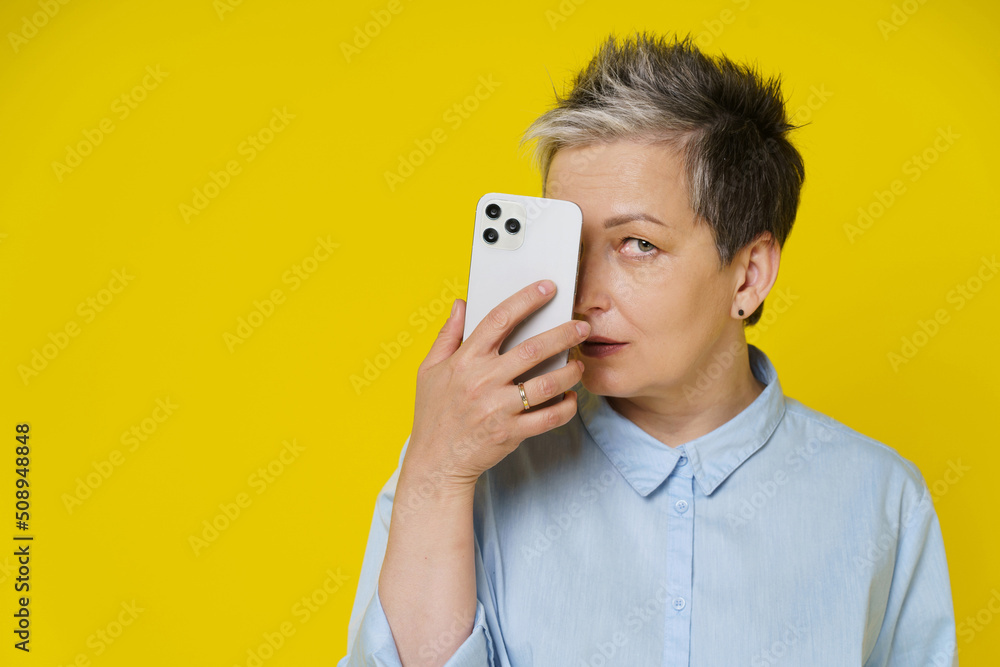 Mature grey haired woman hide face smiling shy behind smartphone or modern gadget working or shopping online or checking on social media. Pretty woman in blue blouse isolated on yellow background Mature grey haired woman hide face smiling shy behind smartphone or modern gadget working or shopping online or checking on social media. Pretty woman in blue blouse isolated on yellow background