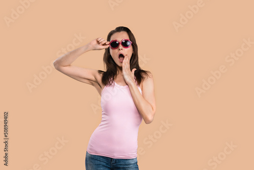 Shocked or wow passionate young woman in sunglasses kiss shaped gesturing oh my god, wearing sleeveless t-shirt posing in studio isolated on pink background © Svyatoslav Lypynskyy