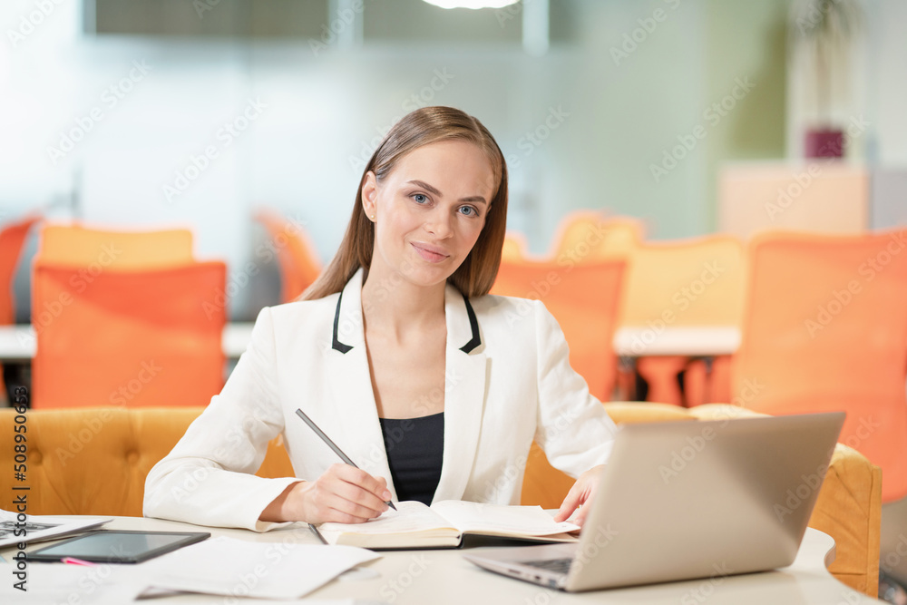 Young business woman in white jacket or suit at the desk with a laptop, looking at camera and making notes. Blonde model female with blue eyes posing in business suit in the office. Business concept Young business woman in white jacket or suit at the desk with a laptop, looking at camera and making notes. Blonde model female with blue eyes posing in business suit in the office. Business concept