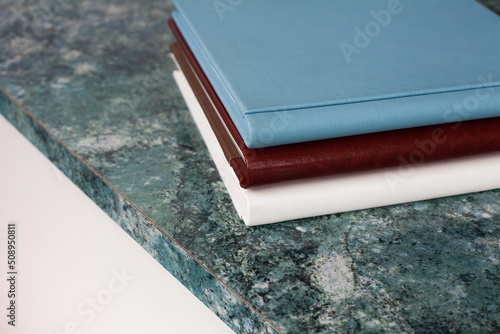 Three stylish photobooks with leather covers, white, burgundy and blue, of different thicknesses, lie on a textured blue surface in a room.