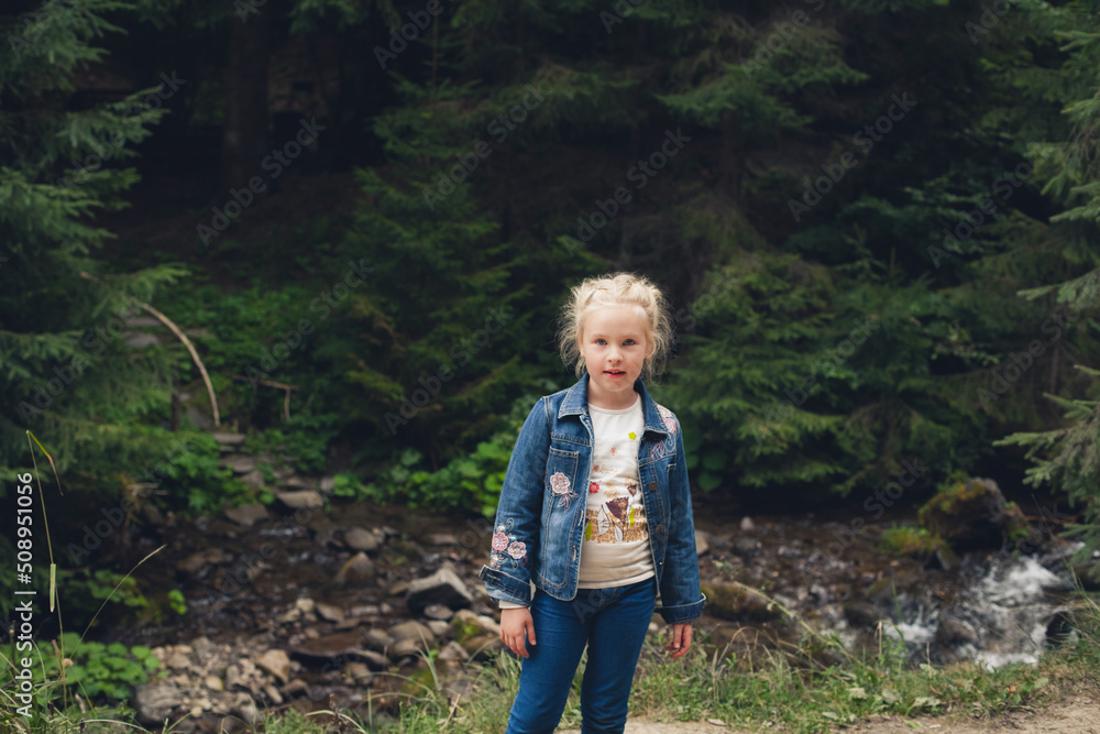 A little girl with blond hair, in jeans clothes, stands against the backdrop of a mountain river with stones and a forest, in summer. A little girl with blond hair, in jeans clothes, stands against the backdrop of a mountain river with stones and a forest, in summer.