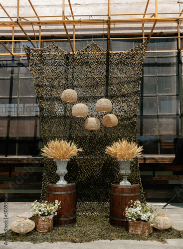 Wedding ceremony in a military style with wicker lamps, baskets with daisies, vases with dry wheat on wooden barrels