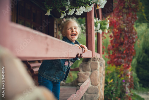 Little cute girl with blond hair laughs. Dressed in a denim suit, she stands at the wooden railing on the veranda of the cottage, decorated with flowers and vegetation. photo