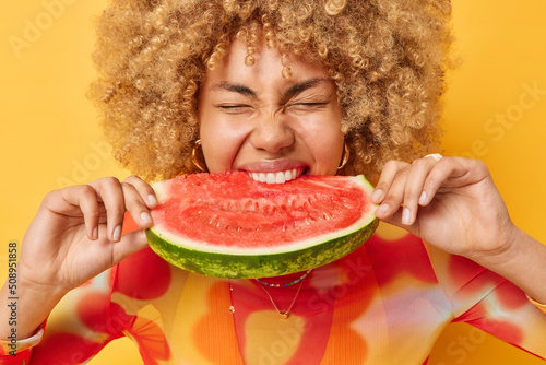 Photo of curly haired young woman eats juicy delicious watermelon bites favorite fruit keeps eyes closed from enjoyment poses against vivid yellow background. Summer time and nutrition concept photo