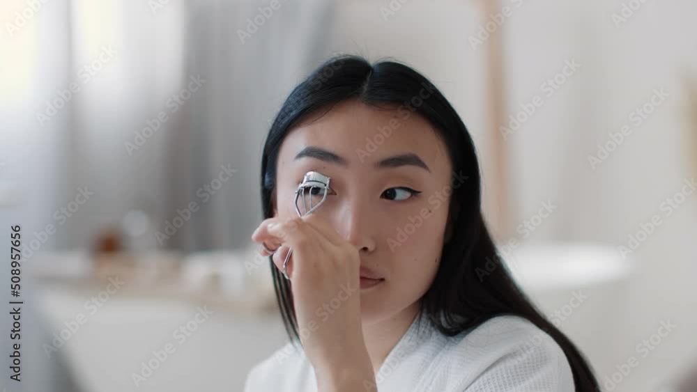 Beauty lifehacks. Young pretty asian woman curling her eyelashes with special curler, looking at mirror at bathroom Beauty lifehacks. Young pretty asian woman curling her eyelashes with special curler, looking at mirror at bathroom
