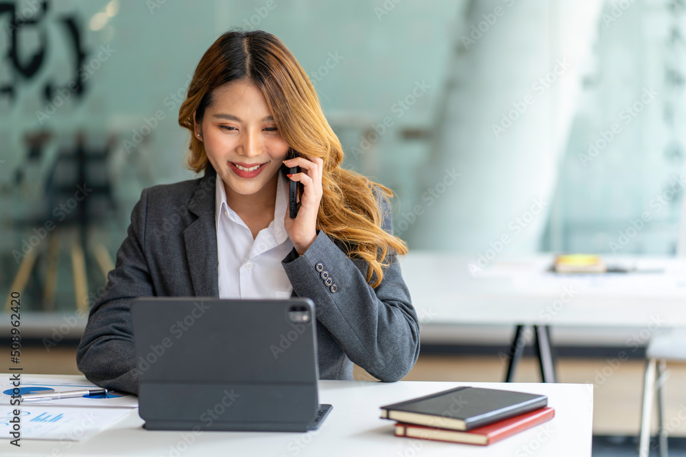 Happy business asian manageress working at her desk in the office taking a call on her mobile phone while writing notes on a notepad Happy business asian manageress working at her desk in the office taking a call on her mobile phone while writing notes on a notepad